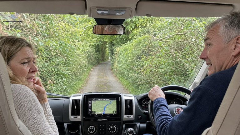 A couple discussing a sat-nav route in a motorhome cab on a narrow lane.
